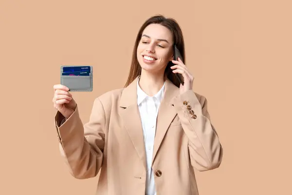 Young woman with credit card talking by mobile phone on brown background