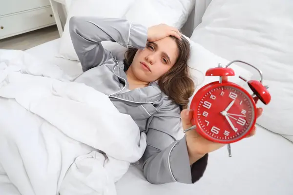 Sleepy young woman with alarm clock lying in bedroom