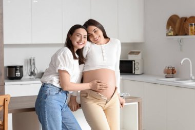 Genç lezbiyen kadın mutfakta hamile karısının karnına dokunuyor.