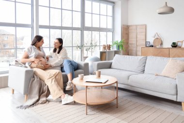 Evdeki kanepede hamile karısının karnına dokunan genç lezbiyen kadın.