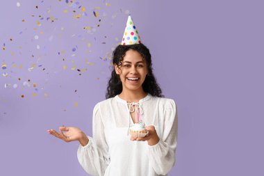 Parti şapkalı güzel Afro-Amerikalı kadın elinde yanan mum ve leylak arkaplanda konfeti olan bir kek tutuyor.