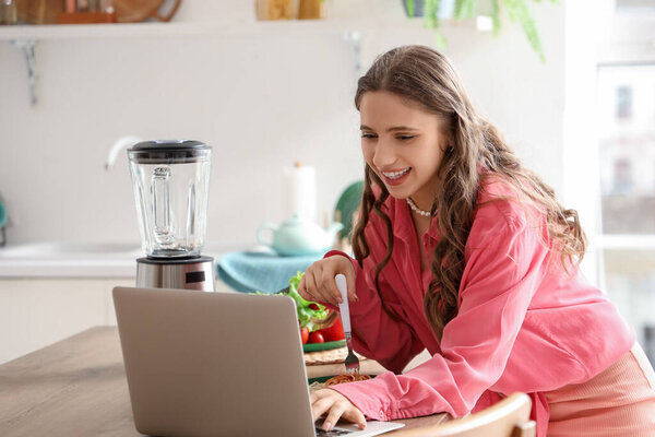 Young woman with laptop on table eating tasty pasta in kitchen