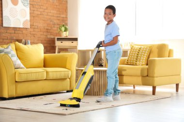 Oturma odasında elektrikli süpürgeyle halı temizleyen yakışıklı Afro-Amerikalı çocuk.