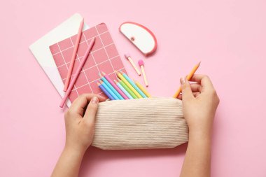 Kalem kutulu çocuk elleri, farklı okul kağıtları ve pembe arkaplan üzerine not defterleri.