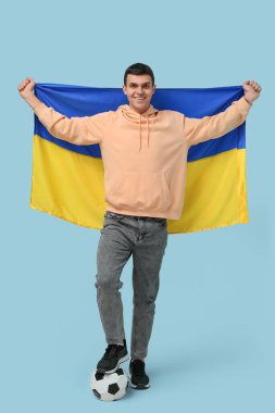 Futbol topu ve Ukrayna bayrağı taşıyan genç adam mavi duvarın yanında.