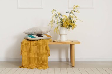 Yumuşak yastıklı ahşap masa, sarı battaniye ve beyaz duvarın yanında mimoza çiçekleri.