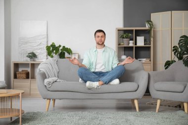 Genç adam evdeki gri kanepede meditasyon yapıyor.