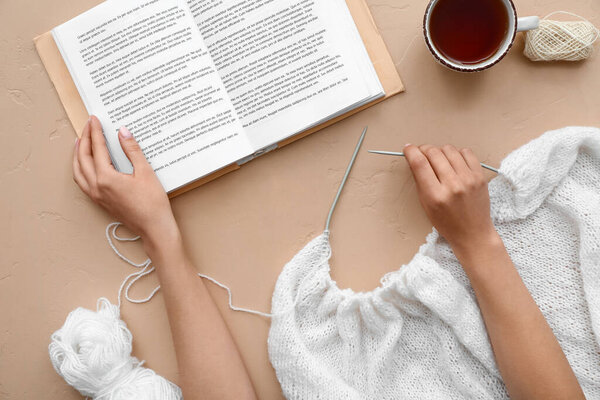 Female hands with knitting needles, book and cup of tea on beige background