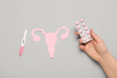 Gri arka planda kağıt uterusu, hapları ve hamilelik testi olan bir kadın eli.