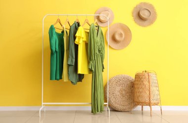 Sarı duvarda şık kadın kıyafetleri ve hasır şapkalar asılı.