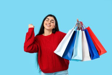 Alışveriş torbalarıyla gülümseyen mutlu genç kadın mavi arka planda evet işareti yapıyor.
