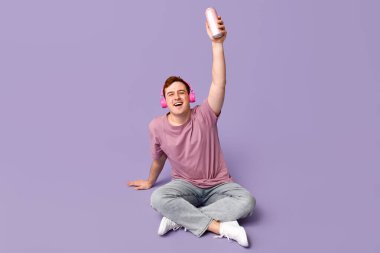 Kulaklıklı genç adam bir kutu sodayla leylak arka planda oturuyor.