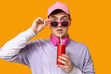 Şok olmuş güneş gözlüklü genç adam sarı arka planda soda içiyor.