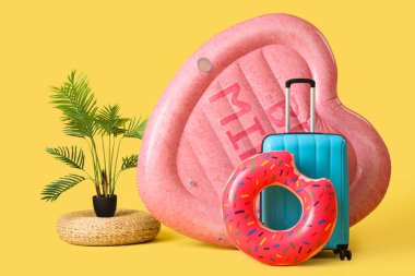 Yüzen kalp şeklinde bir yatak, şişirilebilir yüzük ve sarı arka planda bavul.