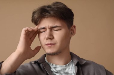 Baş ağrısı olan genç bir adam kahverengi arka planda parmaklarını şakağa bastırıyor, yakın plan.