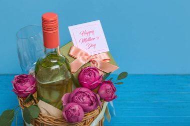 Bir şişe şarap, cam, hediye kutusu ve Anneler Günü için gül çiçekleriyle dolu sepet renkli ahşap masada.
