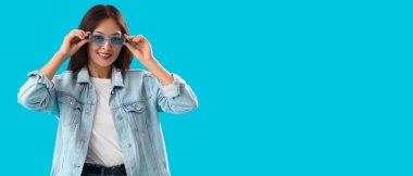 Güneş gözlüklü ve mavi arka planda kot ceketli şık bir Asyalı kadın.