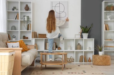 Oturma odasındaki ışık duvarına poster asan genç bir kadın.