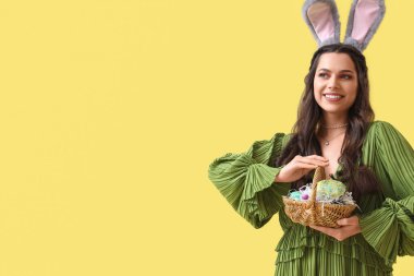 Sarı arka planda Paskalya sepeti olan tavşan kulaklı güzel bir kadın.