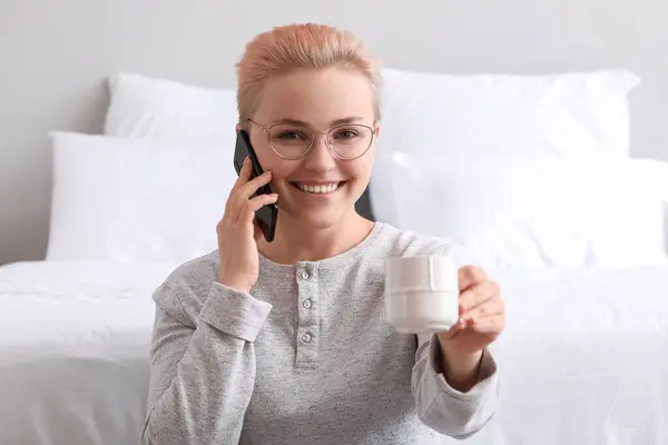 Young woman with cup of coffee talking by mobile phone in bedroom, closeup