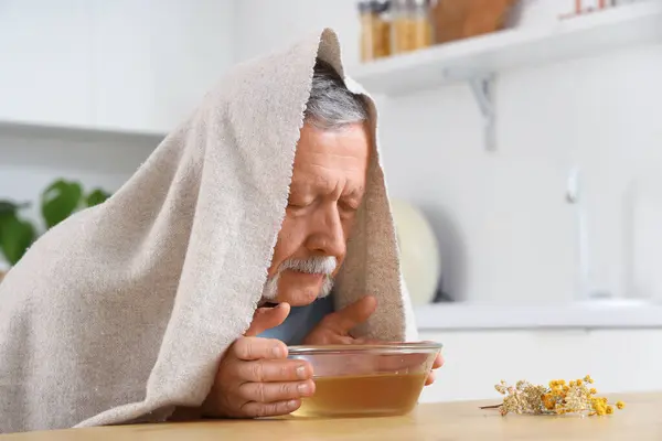 stock image Mature man with towel doing steam inhalation at table in kitchen