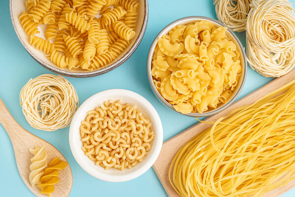 Bowls with different uncooked pasta on blue background