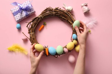 Kadın pembe arka planda boyanmış yumurta, havuç ve tüylerden Paskalya çelengi yapıyor.
