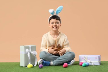 Tavşan kulaklı şirin çocuk. Çikolatalı yumurta sepeti ve paskalya için hediye kutularıyla bej arka planda çimlerin üzerinde oturuyor.