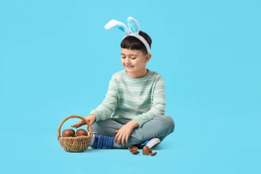 Paskalya için çikolata yumurtası sepeti olan tavşan kulaklı şirin bir çocuk mavi arka planda oturuyor.