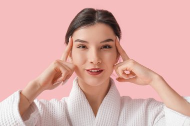 Bornozlu güzel bir kadın pembe arka planda yüz geliştirme egzersizi yapıyor.