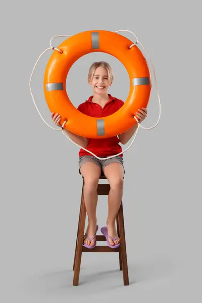 Happy little boy lifeguard with ring buoy sitting on chair against grey background