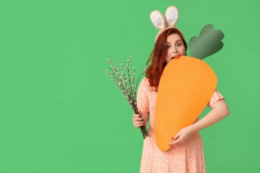 Beautiful young woman in bunny ears with pussy willow branches and paper carrot on green background. Easter celebration