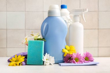 Beyaz masadaki temizlik malzemeleri ve bahar çiçekleri fayanslı arka planda.