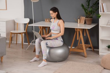 Evde spor yaparken dizüstü bilgisayar kullanan genç bir kadın.