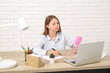 Elinde mini vantilatör olan güzel genç bir kadın ofiste çalışıyor.