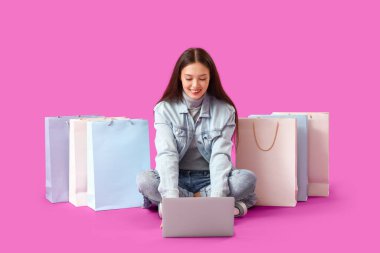 Dizüstü bilgisayarı ve mor arka planda alışveriş torbaları olan genç Asyalı kadın.