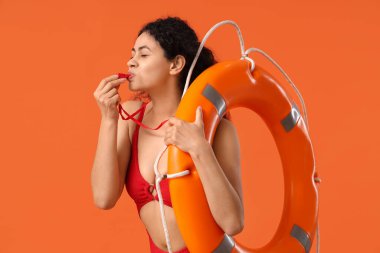 Beautiful young African-American female lifeguard with ring buoy whistling on orange background clipart