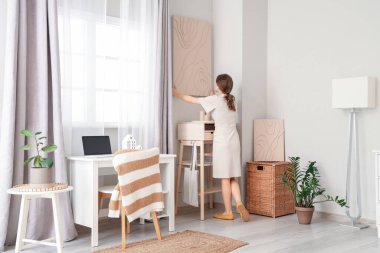 Çalışma odasının yakınındaki duvara resim asan genç ve güzel bir kadın.