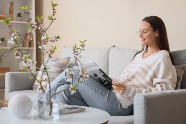 Şık oturma odasındaki kanepede çiçek açan dallarla dergi okuyan genç bir kadın.