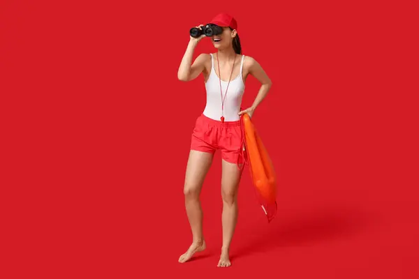 Female lifeguard with rescue tube buoy looking through binoculars on red background