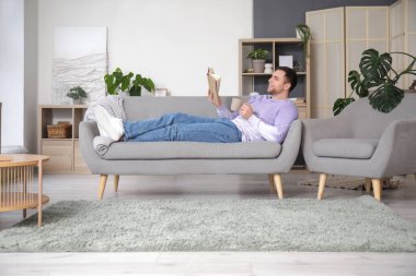 Evdeki gri kanepede çay fincanı okuyan genç adam.