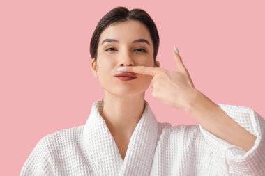 Bornozlu güzel bir kadın pembe arka planda yüz geliştirme egzersizi yapıyor.