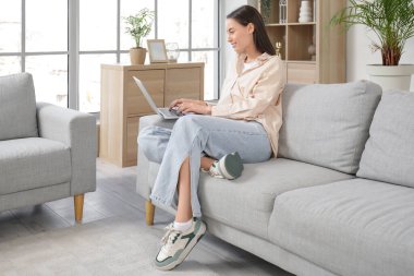 Oturma odasındaki gri koltukta dizüstü bilgisayarı olan güzel genç bir kadın oturuyor.