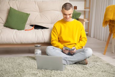 Yerde dizüstü bilgisayarı, kavanoz bankası ve odada parası olan bir erkek öğrenci. Eğitim için tasarruf kavramı