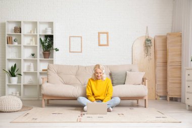 Kulaklık takan olgun bir kadın evde dizüstü bilgisayar kullanıyor.