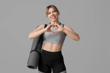 Yoga minderli genç bayan fitness eğitmeni gri arka planda kalp hareketi gösteriyor.
