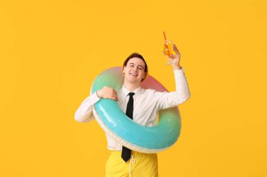 Şişme yüzüklü, kokteylli ve paletli bir ofis çalışanı sarı arka planda tatil hayali kuruyor.