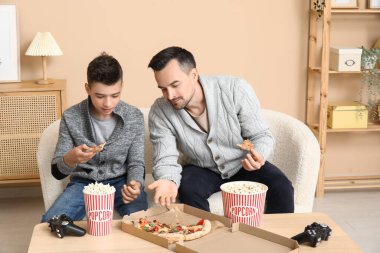 Baba ve küçük oğlu evdeki kanepede lezzetli yemekler yiyorlar.