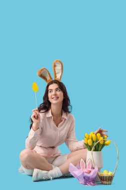 Tavşan kulaklı güzel bir kadın. Paskalya yumurtası, pasta ve laleler mavi arka planda oturuyor.