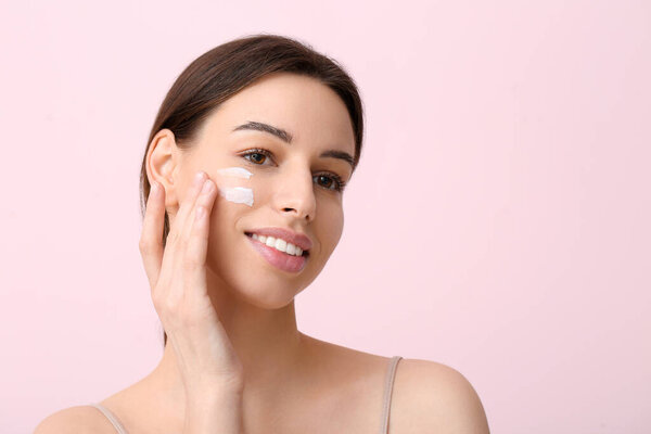 Красивая молодая женщина наносит крем для лица на розовый фон